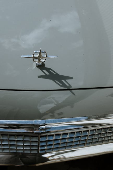 Close-up of a vintage car hood showcasing a chrome emblem casting a shadow, reflecting classic automotive design.