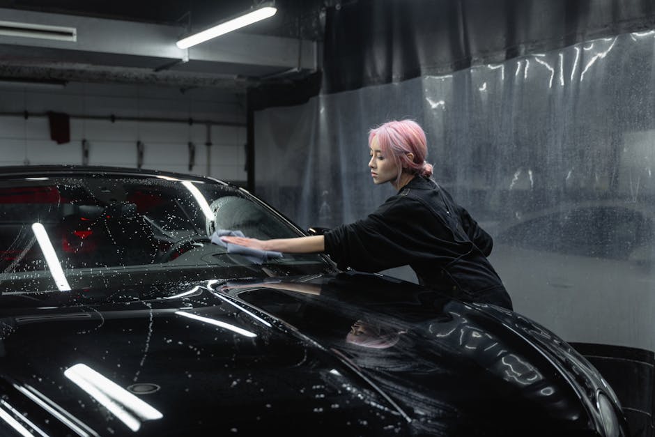 An adult woman with pink hair meticulously wiping down a black car at an indoor car wash.