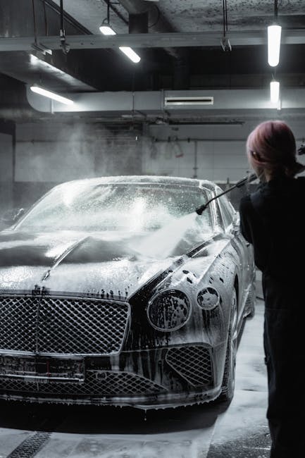 A sleek black luxury car getting washed with foam by a worker in an indoor garage.
