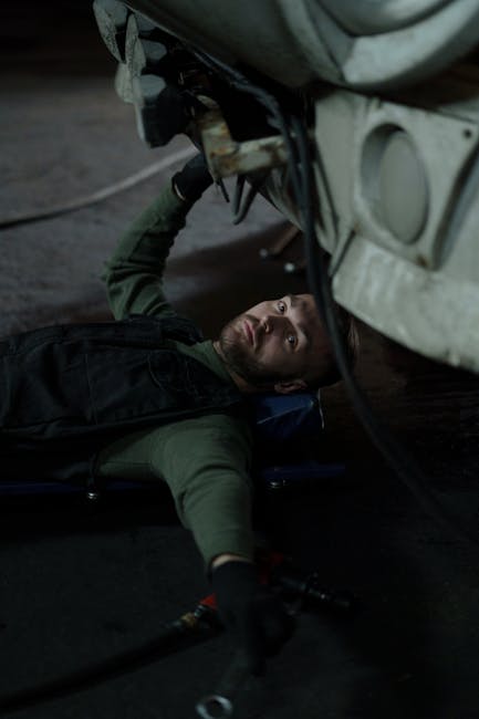 Man in overalls lying on creeper beneath vehicle in dark garage.